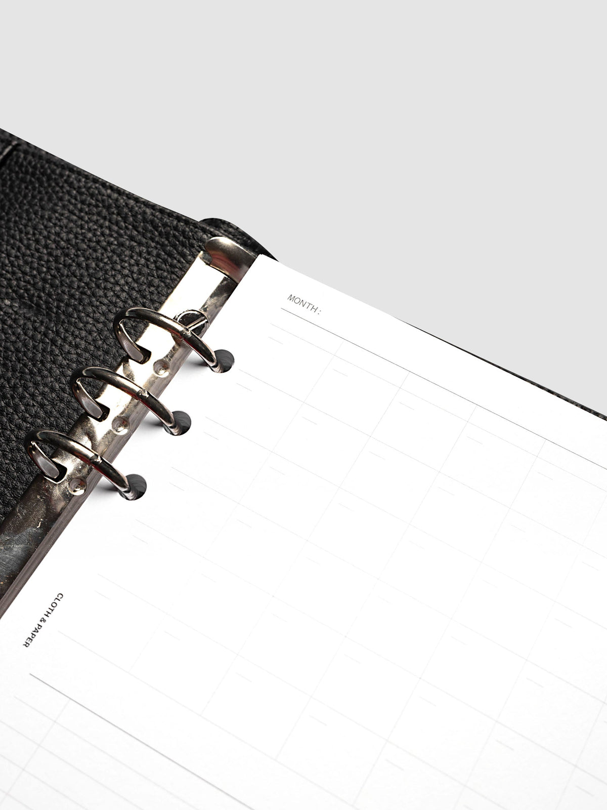 Monthly Fold Out Planner Insert Set of 2, A5 6-Ring Punch, angled close-up of the top left corner showing the month line, the gray calendar grid, and the silver rings against black pebbled leather