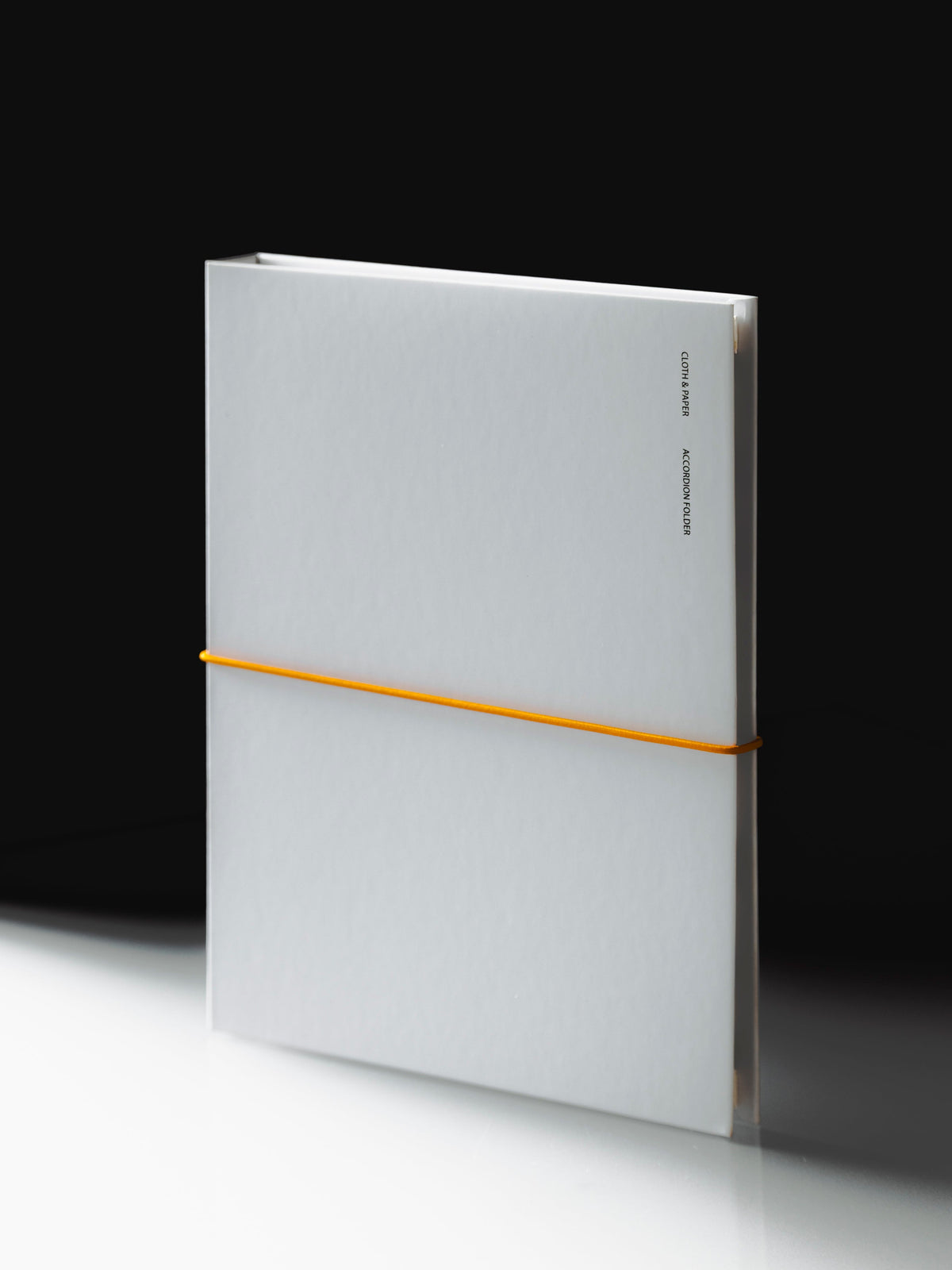 The Editorial Accordion Folder standing closed, a light gray cover held shut with an orange elastic band, on a white surface against a black background.