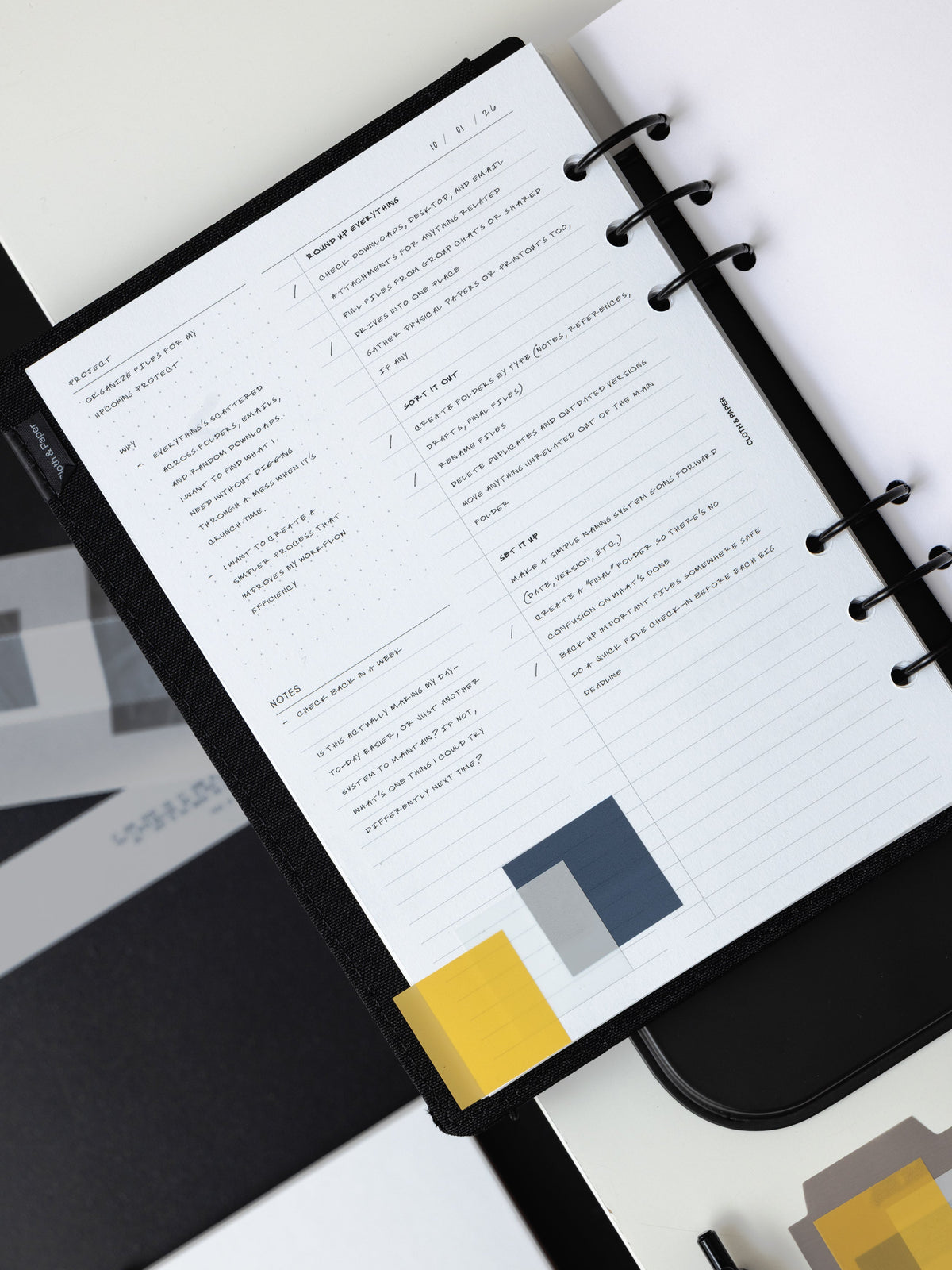 A black six ring planner open to a filled-in planner insert, marked with transparent Square Page Flags in yellow, navy and gray, on a white surface.