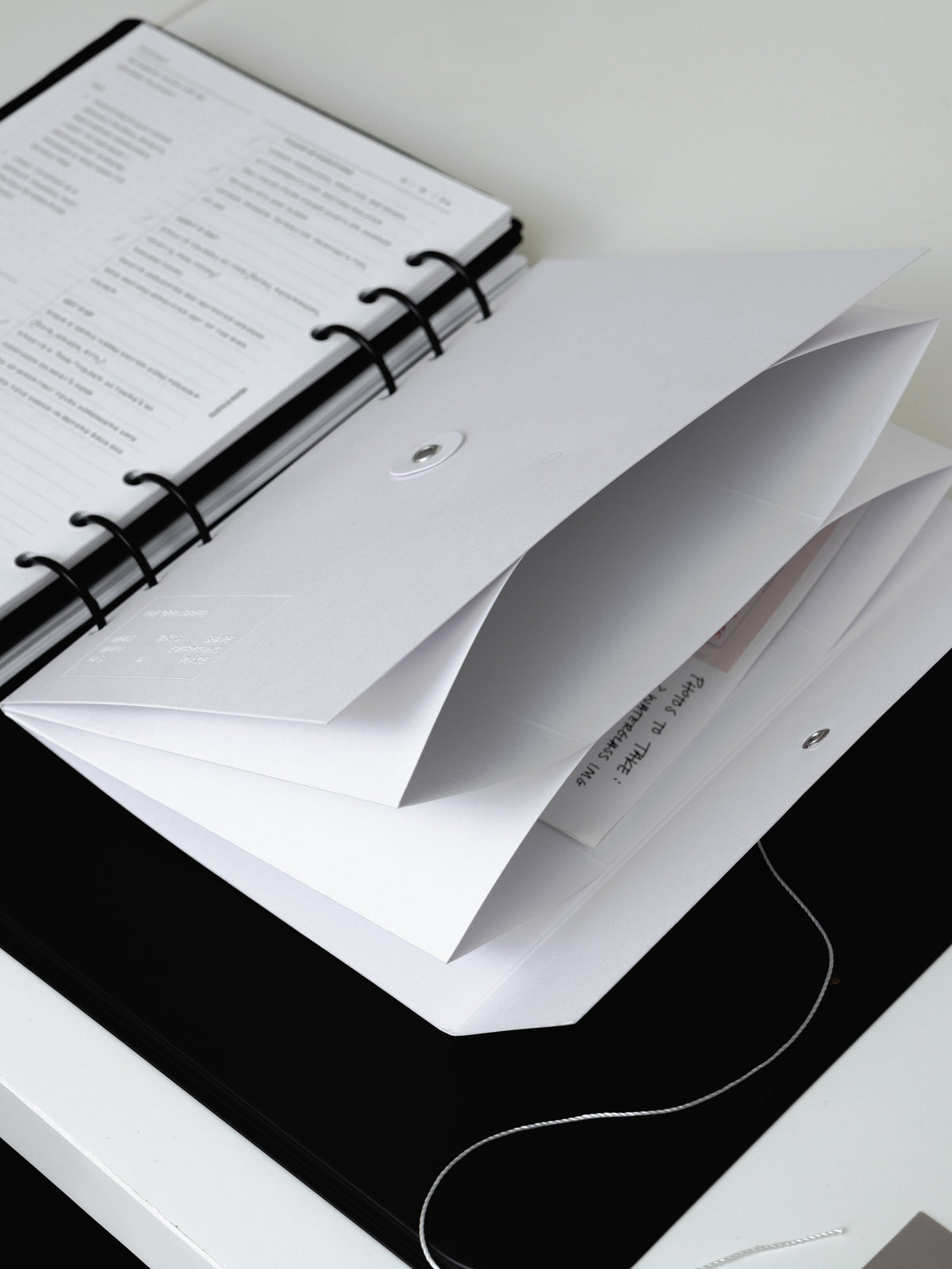 An Open Brief Dashboard in a black six ring planner, its two white folder pockets fanned open and its string closure trailing across a white surface.