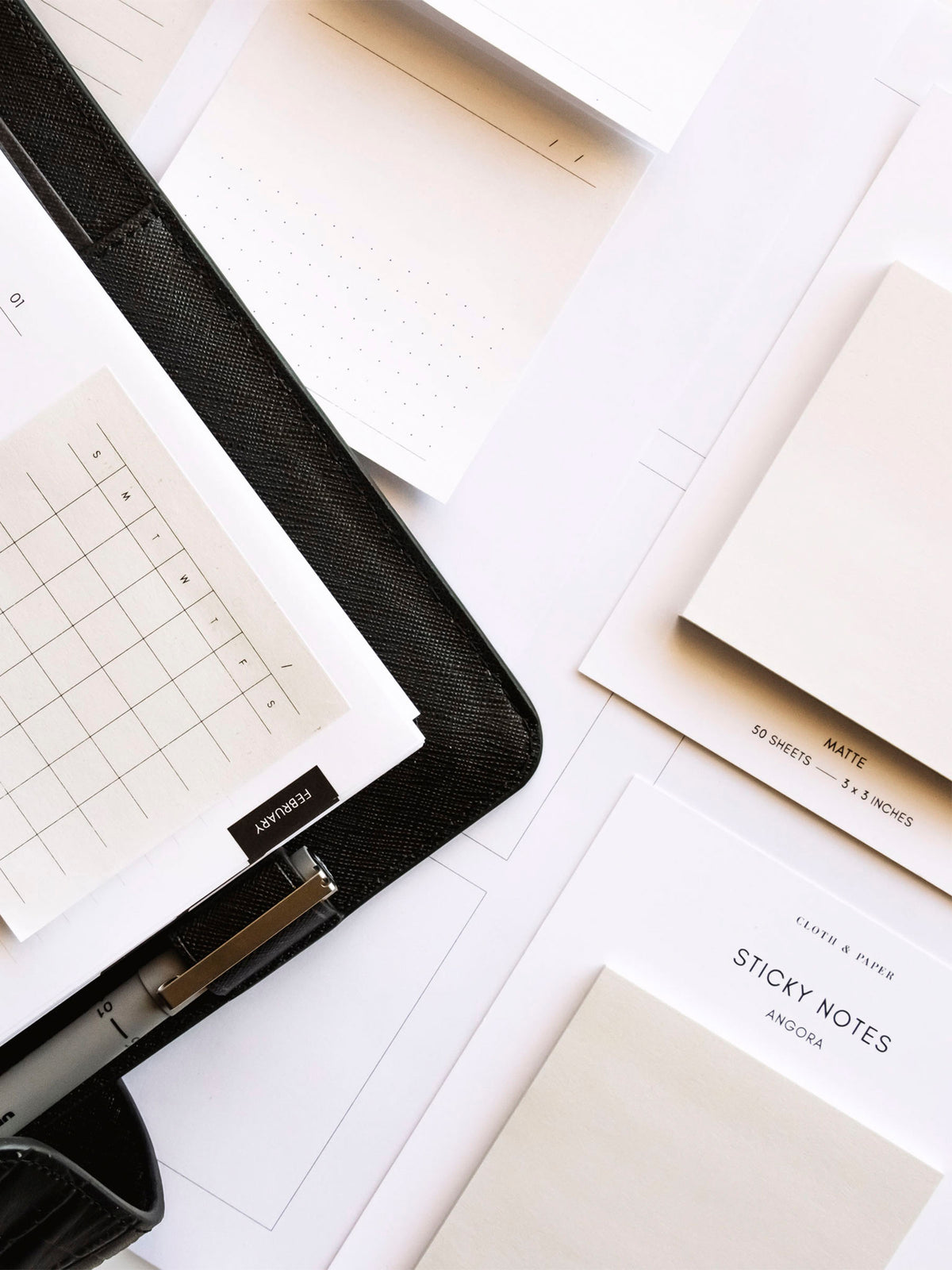 Printed sticky note stuck on the page inside a spiral planner. Spiral planner has a Leather Agenda Cover with a Uni Pin in the cover's pen loop. There are sticky note pads and more printed sticky notes on the table around the planner.
