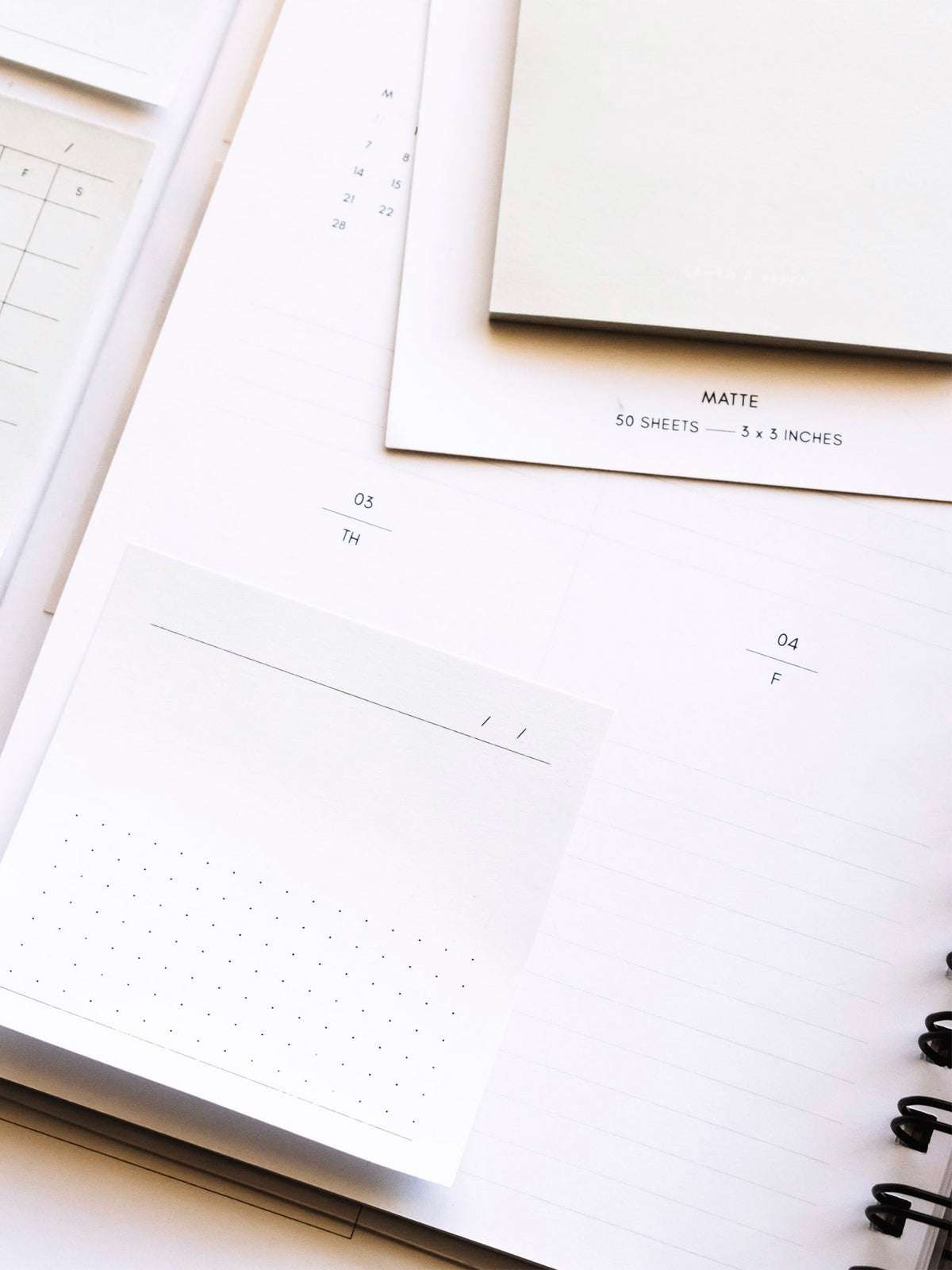 Sticky note printed using the 3x3 Sticky Note Printable in Essentials stuck to the bottom left corner of a spiral bound planner. A pad of sticky notes rest on top of the planner, and the rest of the printed sticky notes rest on the table to the left of the planner.