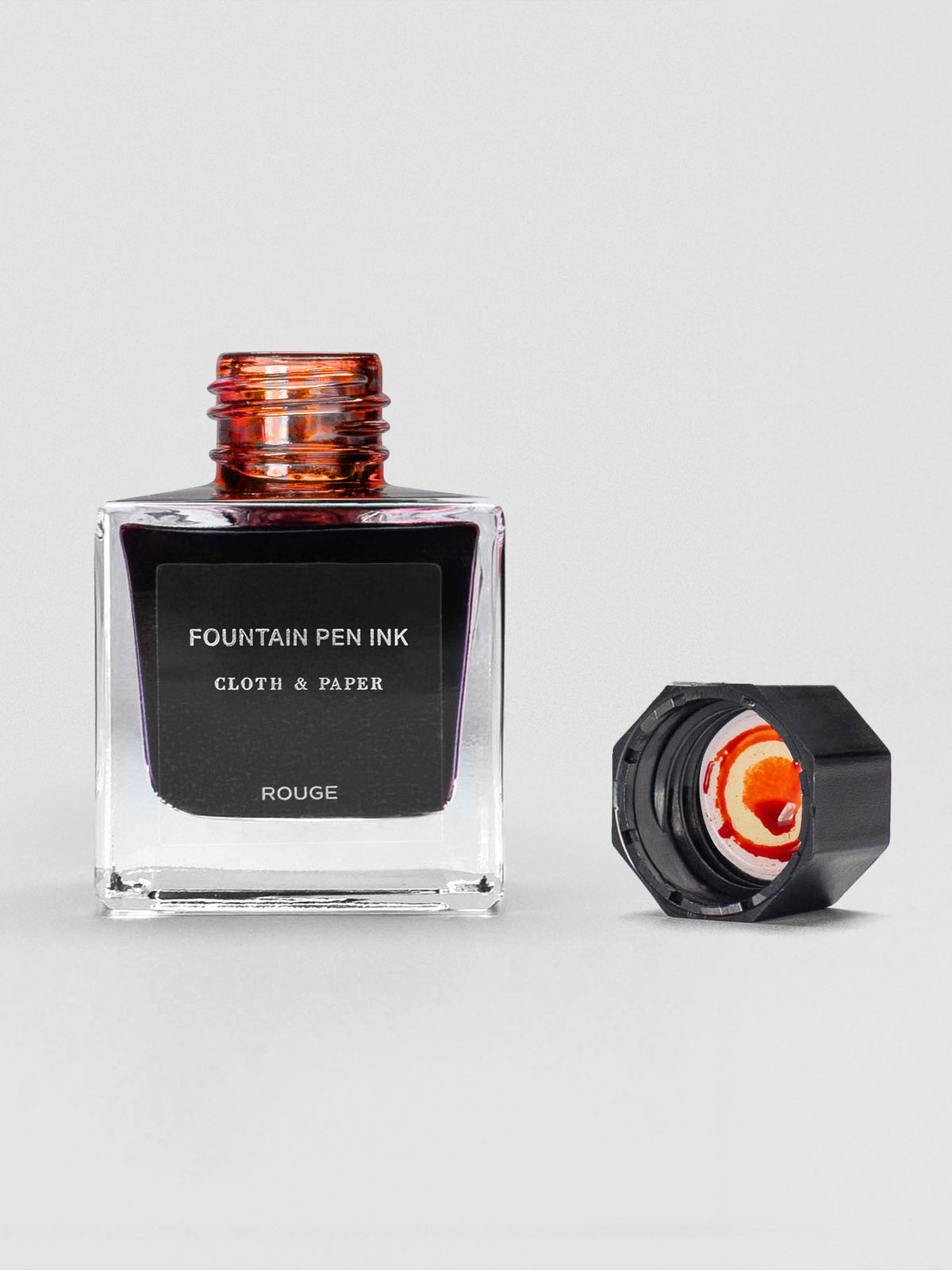 A square glass bottle of Cloth & Paper Fountain Pen Ink in Rouge with the cap removed, highlighting a warm red-orange tint at the bottle opening and ink residue inside the cap.