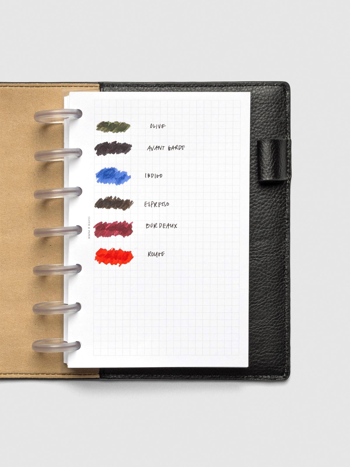 Fountain pen ink swatches on a grid notebook page inside a black leather planner cover. Six ink colors are shown in labeled swatches: Olive (green), Avant Garde (dark brown-black), Indigo (blue), Espresso (warm brown), Bordeaux (deep red), and Rouge (bright red).