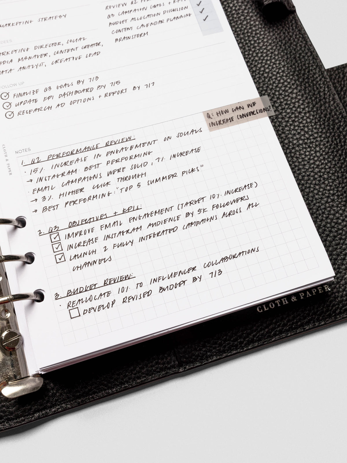 Close-up of a meeting notes insert inside a black leather planner, filled with handwritten agenda points, follow-up tasks, and gridded notes.