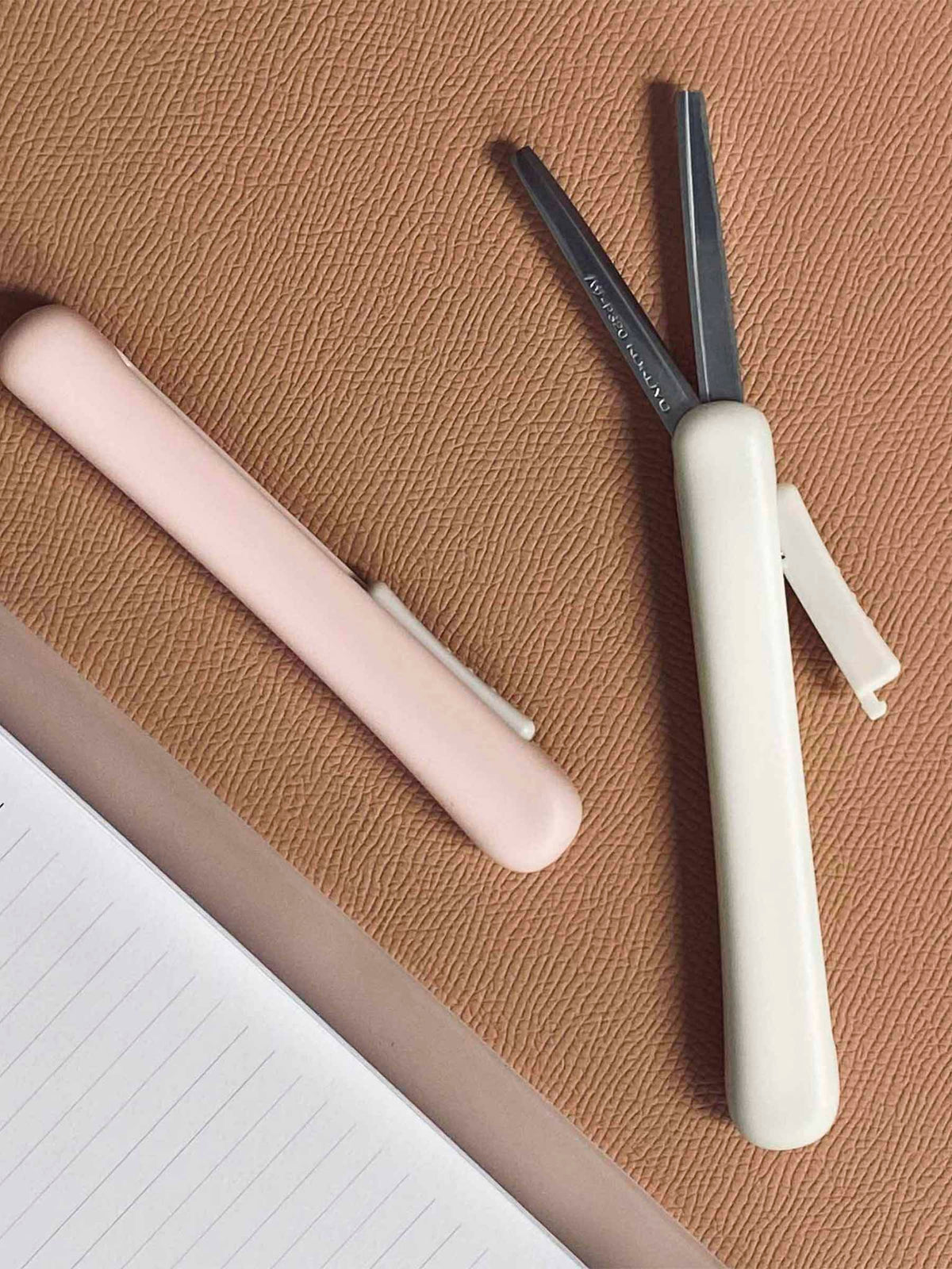 White and Peach scissors on a rust colored surface next to a planner. The white scissors are extended to show the blades.