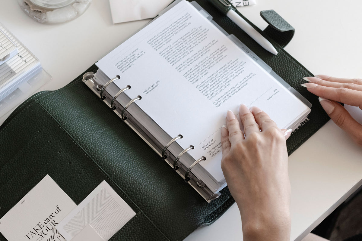 A Foundations Leather 6-Ring Agenda in A5 is opened on a white desk. Two hands are beginning to flip through the pages inside.