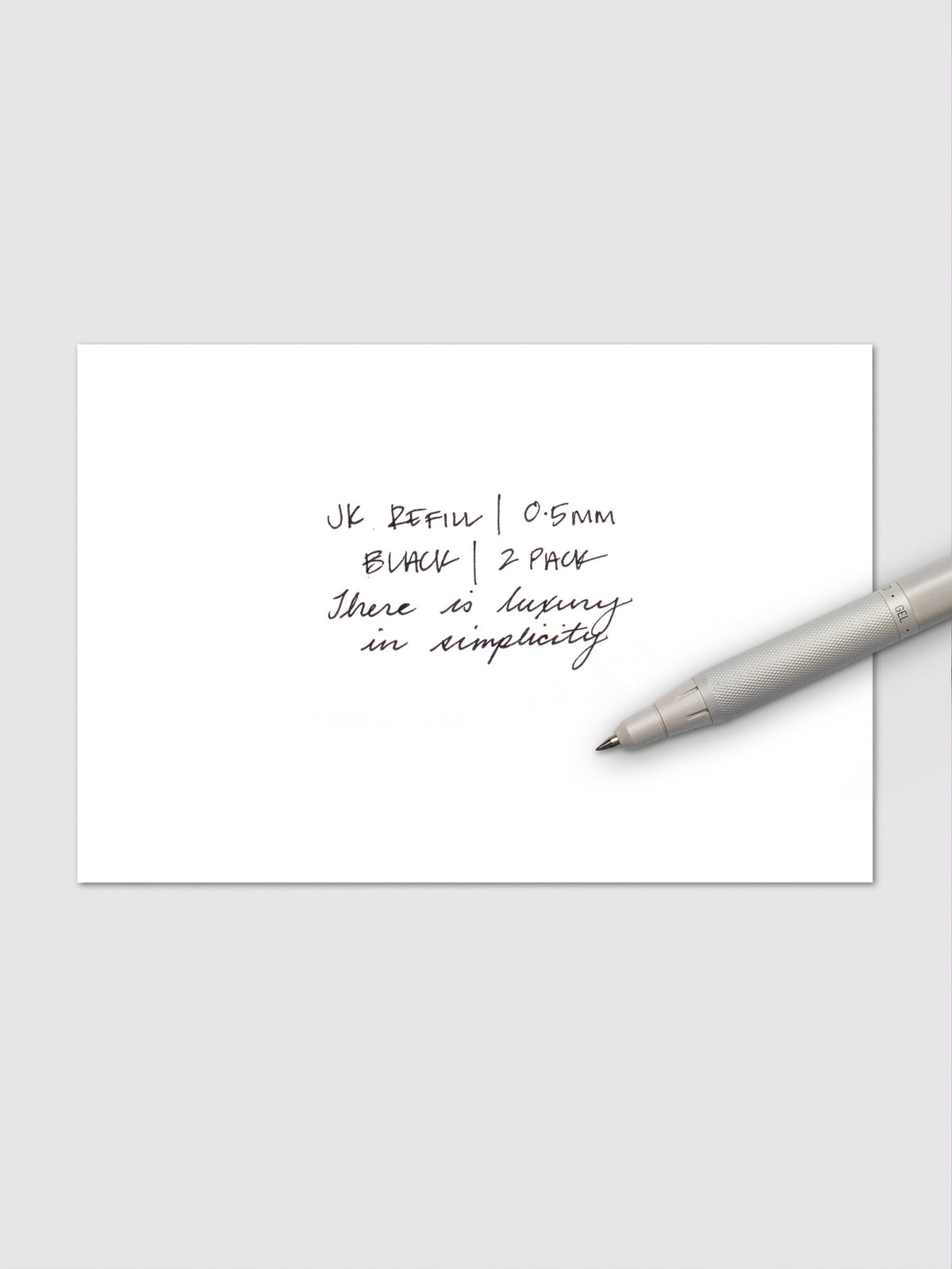 Close-up of handwritten text in black ink reading "JK Refill | 0.5mm Black | 2 Pack" followed by "There is luxury in simplicity," with a white Zebra pen resting on the paper.
