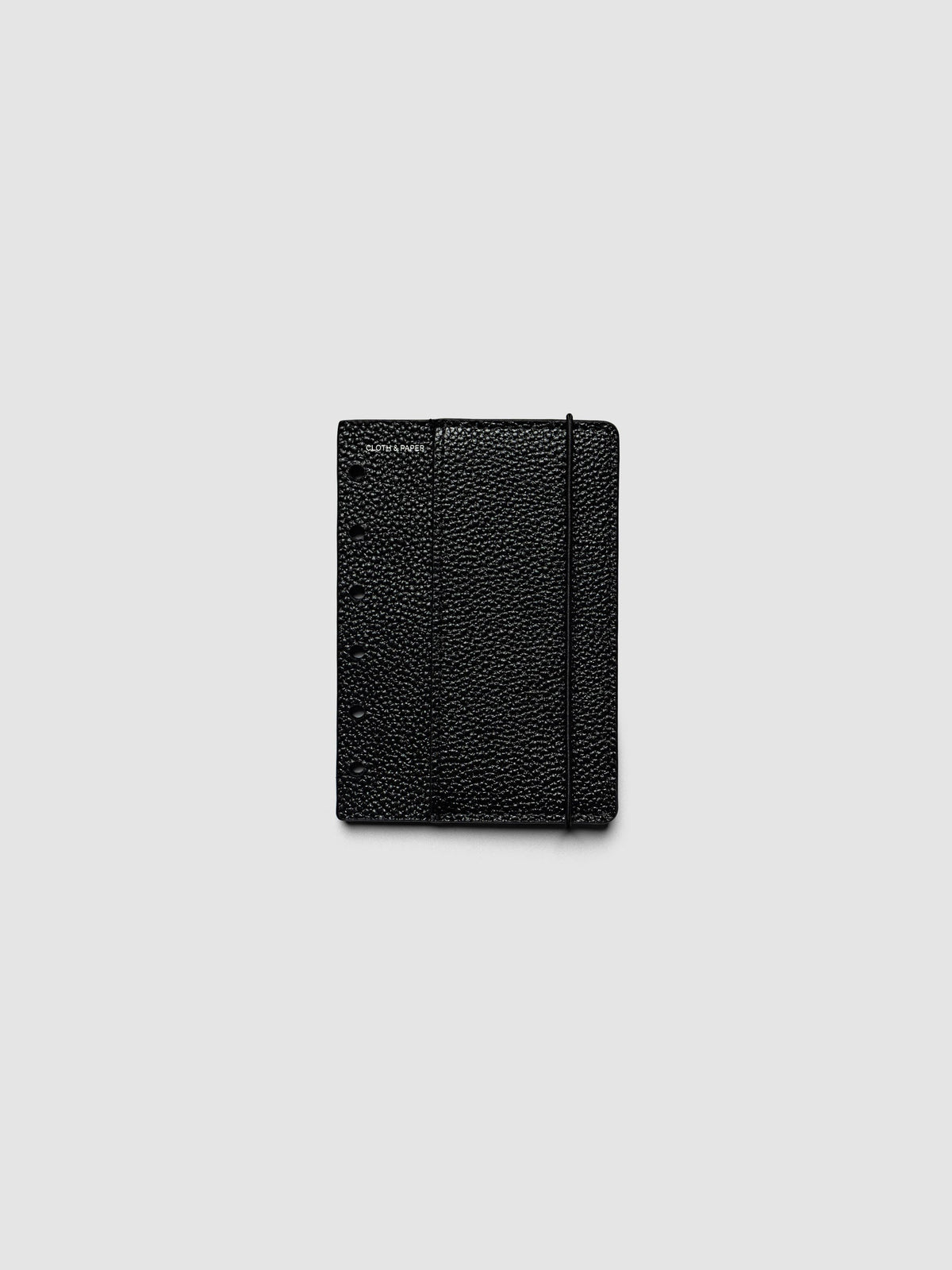 The front cover of the Pocket Genuine Leather Planner Cover Set laid flat, showing pebbled black leather, the silver foiled logo at the top left, the punch holes down the left edge, and the bungee cord running down the right