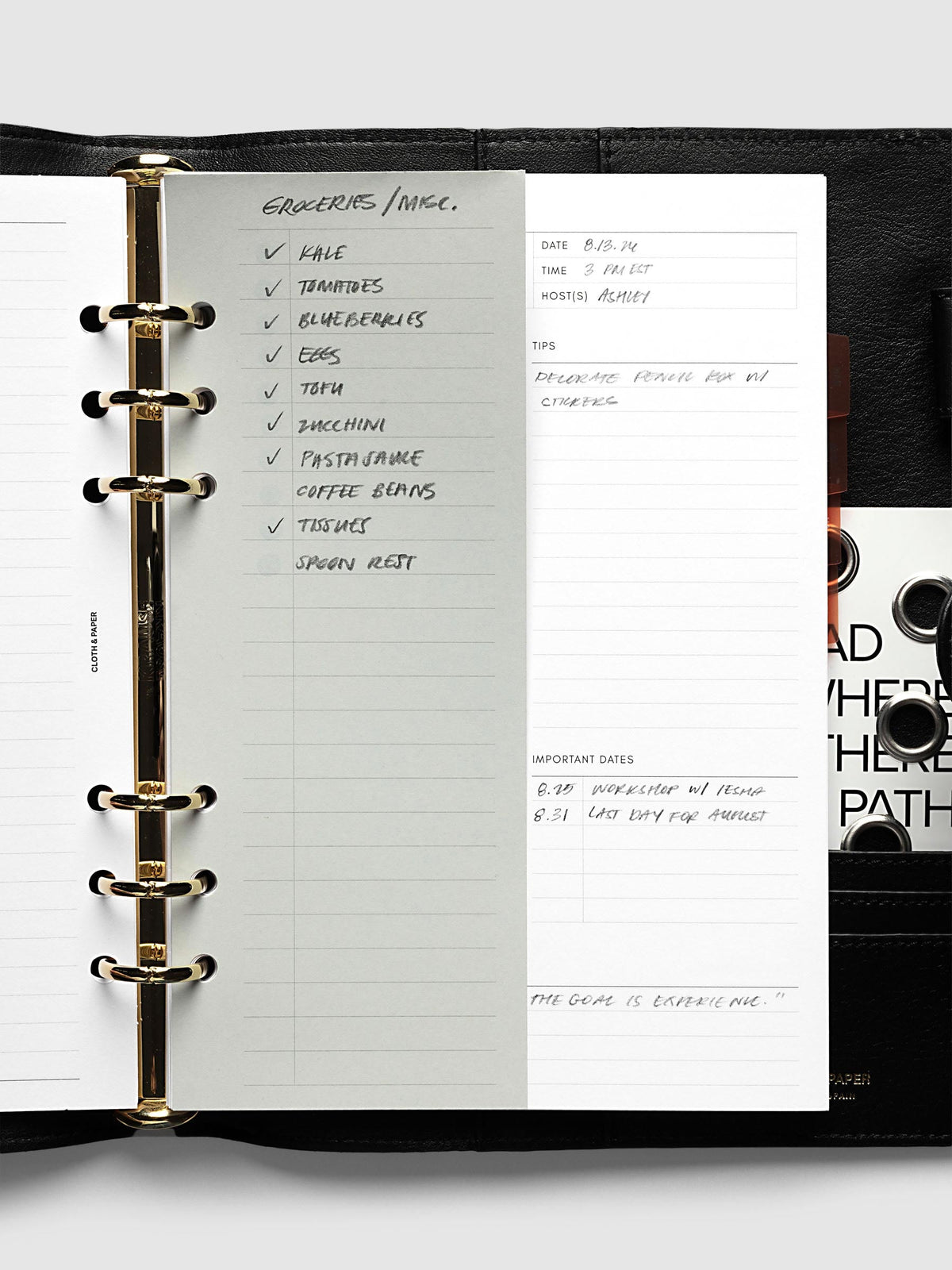 System Punch Pad, A5 6-Ring Punch, sheet clipped onto the gold rings of an open black leather planner, a handwritten grocery list checked off down the page