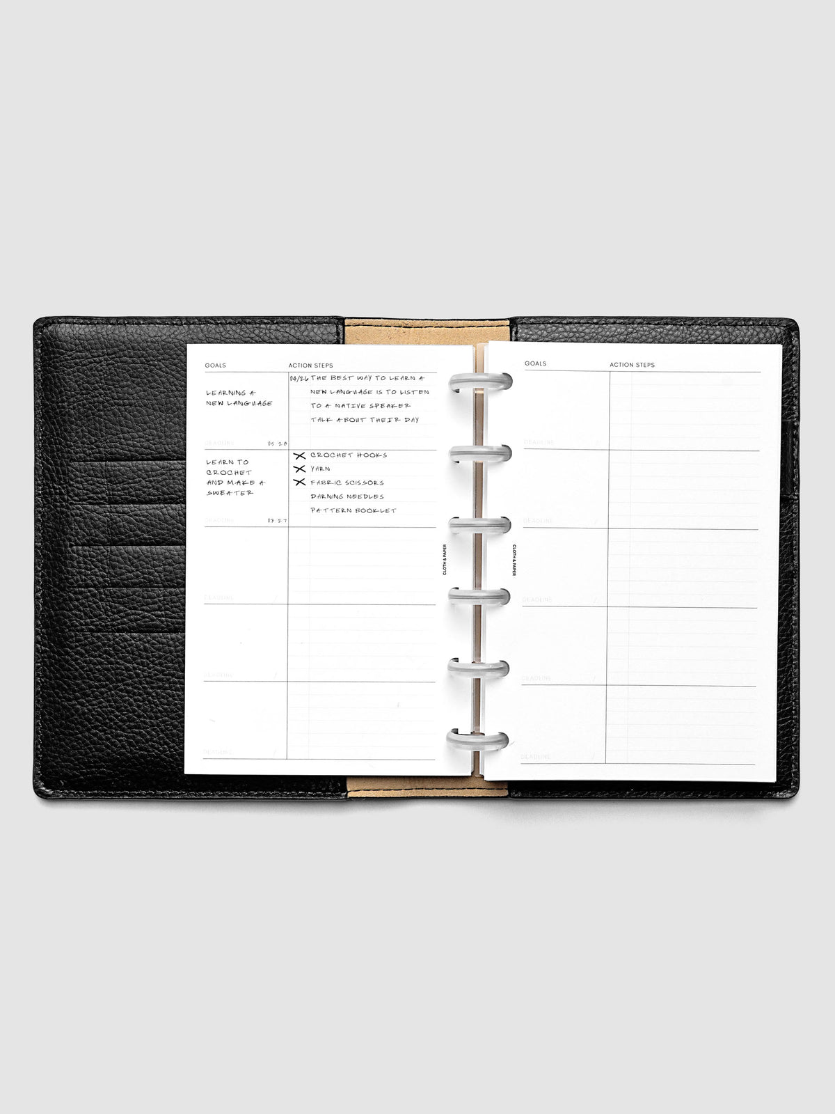 Open black leather planner displaying inserts with weekly goals and action steps page and sample text. The planner is set against a plain light gray background.
