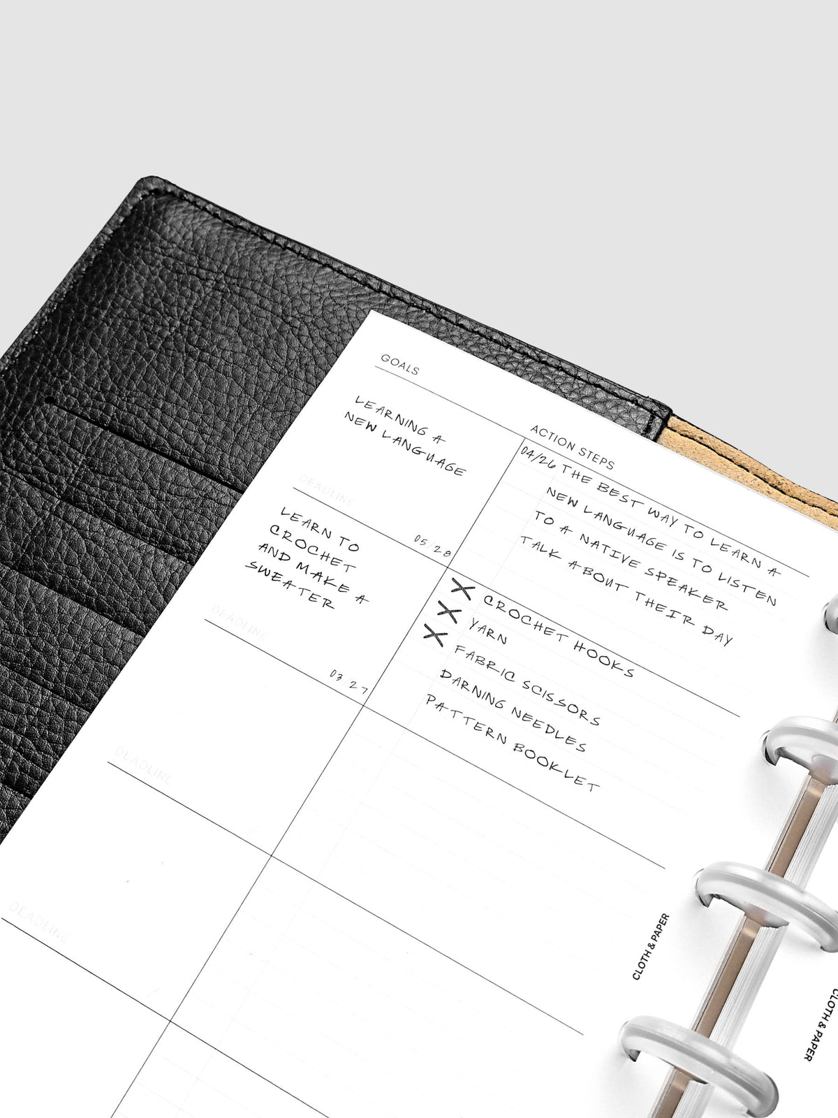 Open black leather planner displaying a goals page with handwritten tasks and action steps, including learning a new language and buying craft supplies. The planner is open on a white surface.