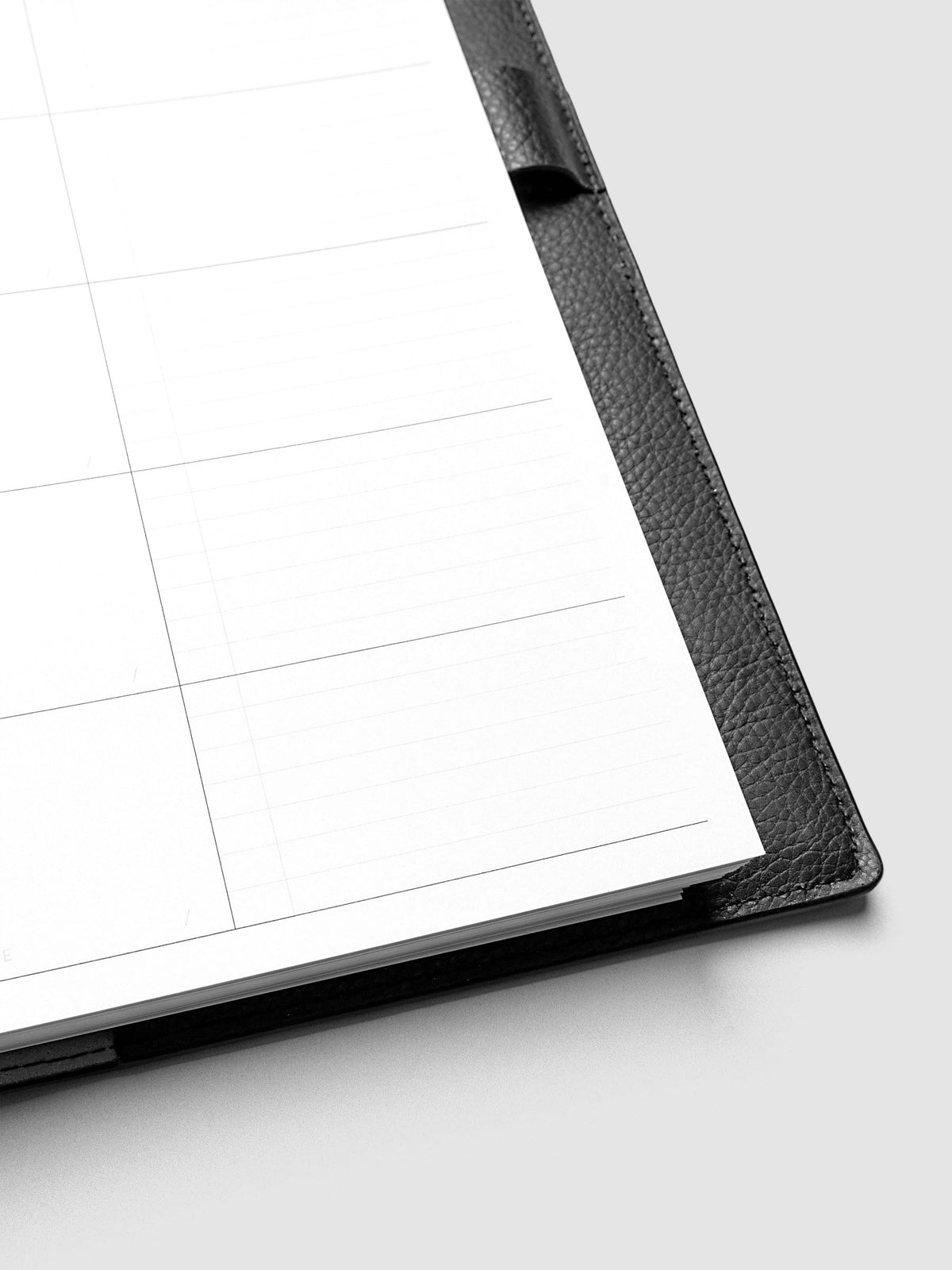 Close-up of an open black leather planner displaying a blank, lined and grid page. The planner rests on a light gray surface.