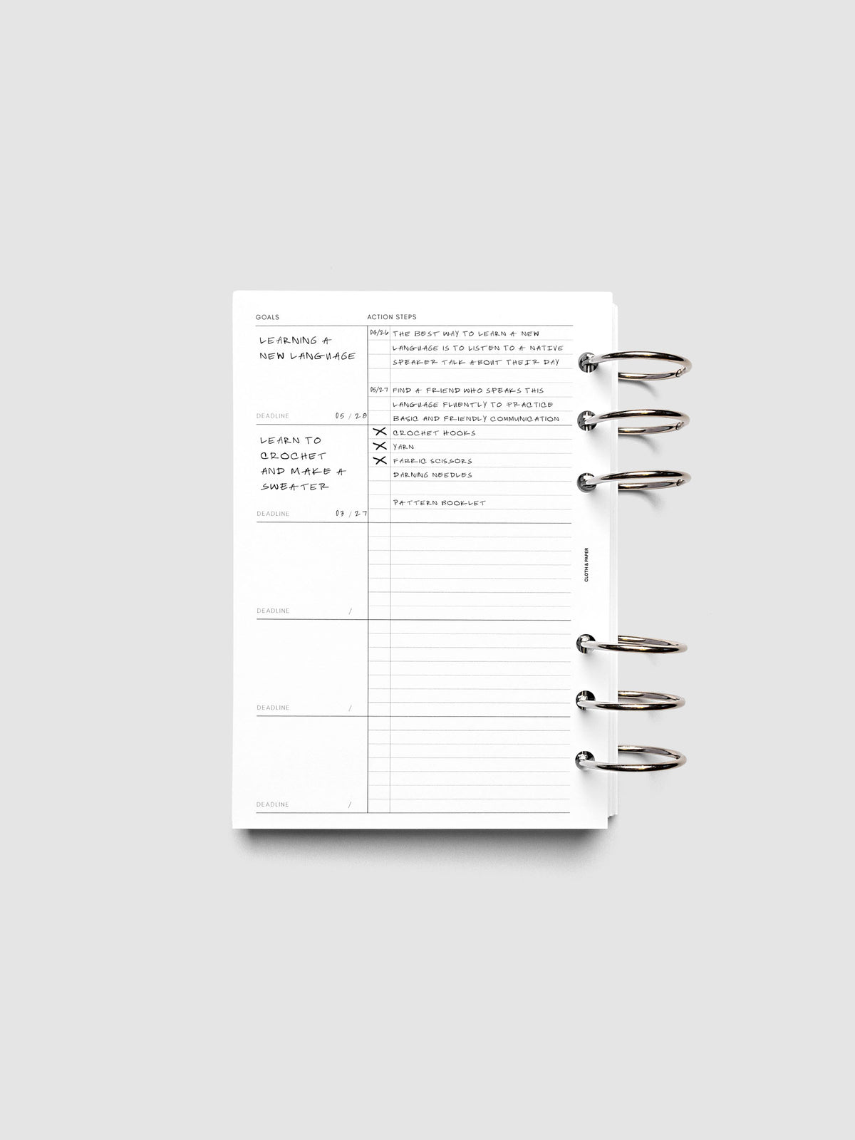 Open ring-bound planner displaying a goals and action list page, with handwritten notes about learning a new language and steps to achieve it. The planner is laid flat on a light gray background.
