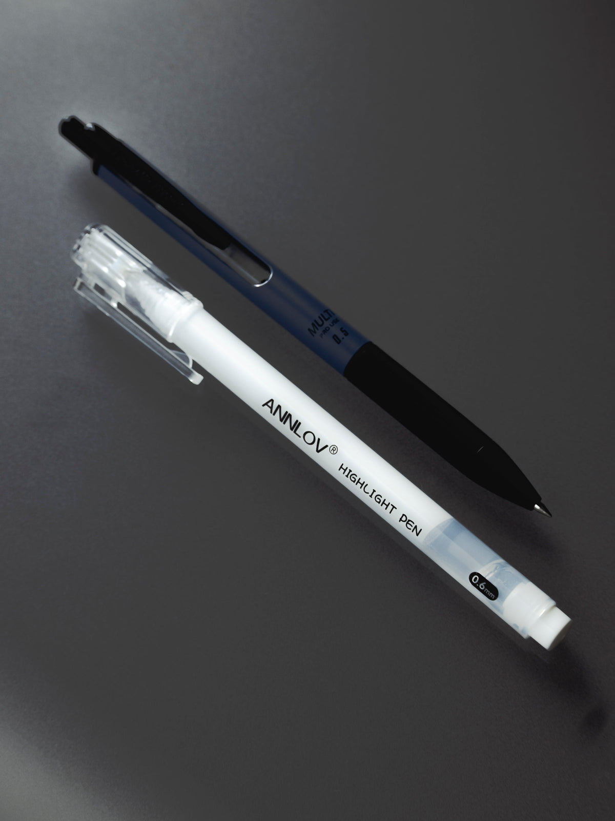 A white Annlov Highlight Pen laid across a Youngmoon Metal Gel Pen in peacock blue and black, on a charcoal surface.