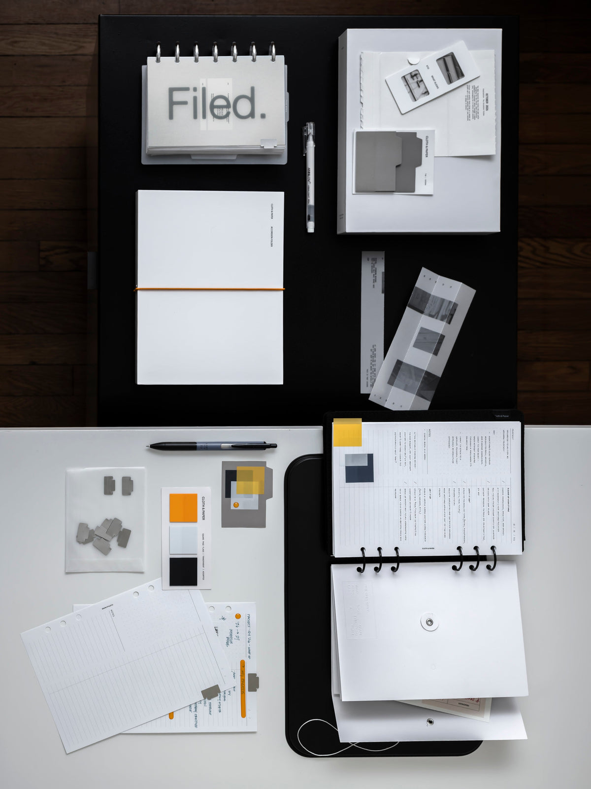 Overhead view of the full October Intention Box on a black and white surface, including the frosted Filed Dashboard Duo on a disc planner, the Editorial Accordion Folder with its orange band, the Accordion Folder Photo Slide, gray Tab Sticky Notes, orange Raised Stickers, two pens, and a black six ring planner.