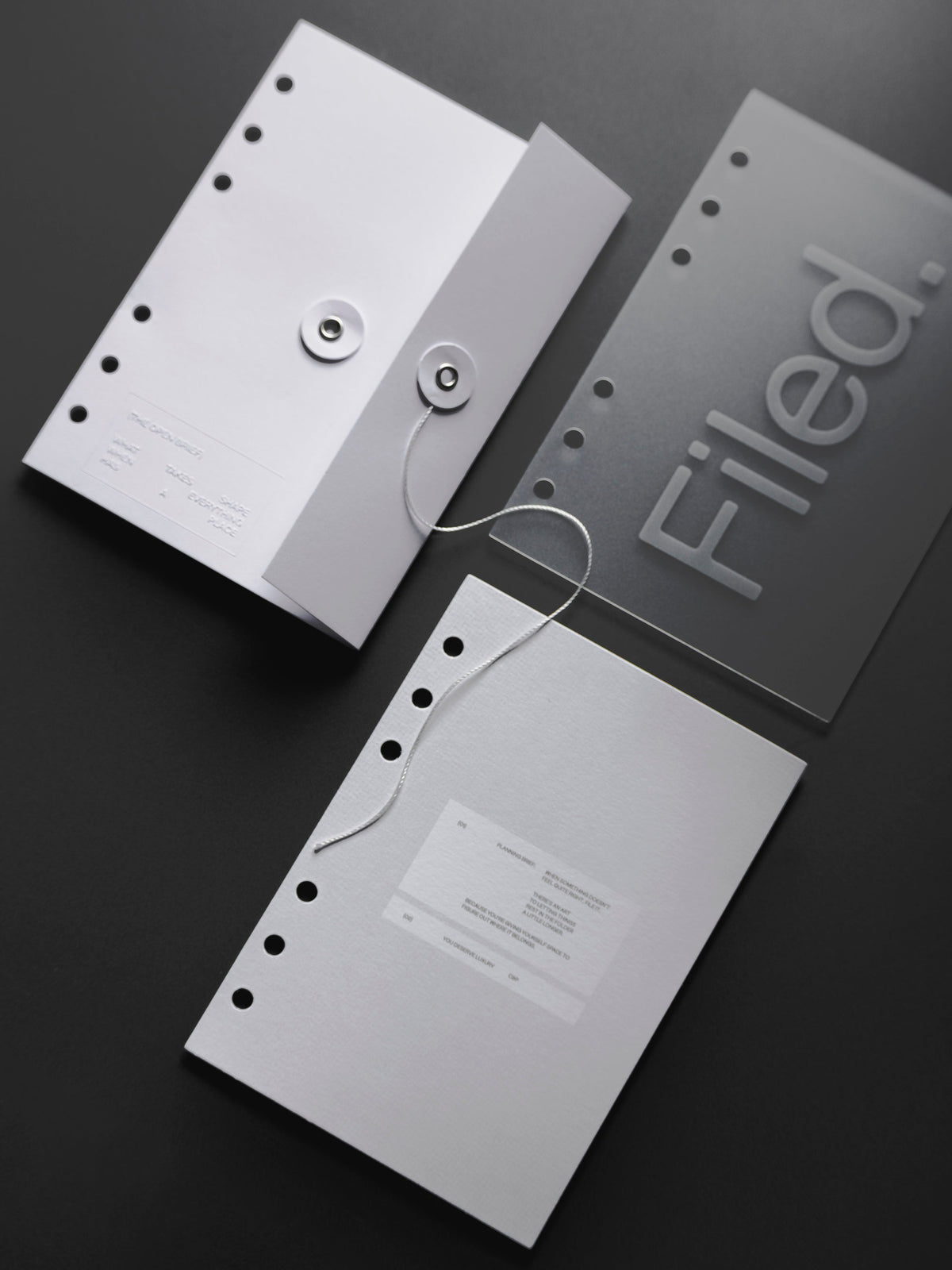 An Open Brief Dashboard with its white string and silver grommet closure beside both layers of the Filed Dashboard Duo, a frosted sheet printed with Filed. in silver and a white card with a text box, all six ring punched, on a charcoal surface.