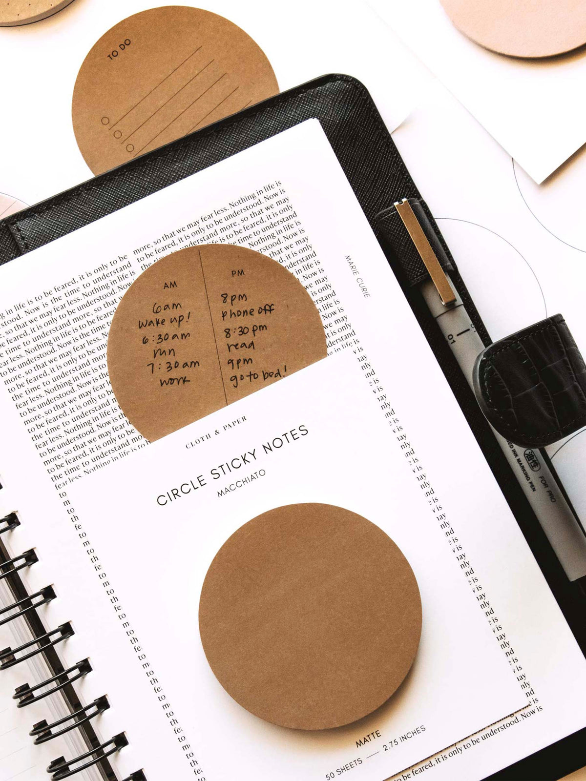 Pad of blank Circle Sticky Notes in Macchiato resting on top of a planner. The planner page shown has a printed, in-use Circle Sticky Note resting on the page. There is a Uni Pin inside the planner's pen loop. Other printed sticky notes and blank sticky note pads are resting on the table underneath the planner.