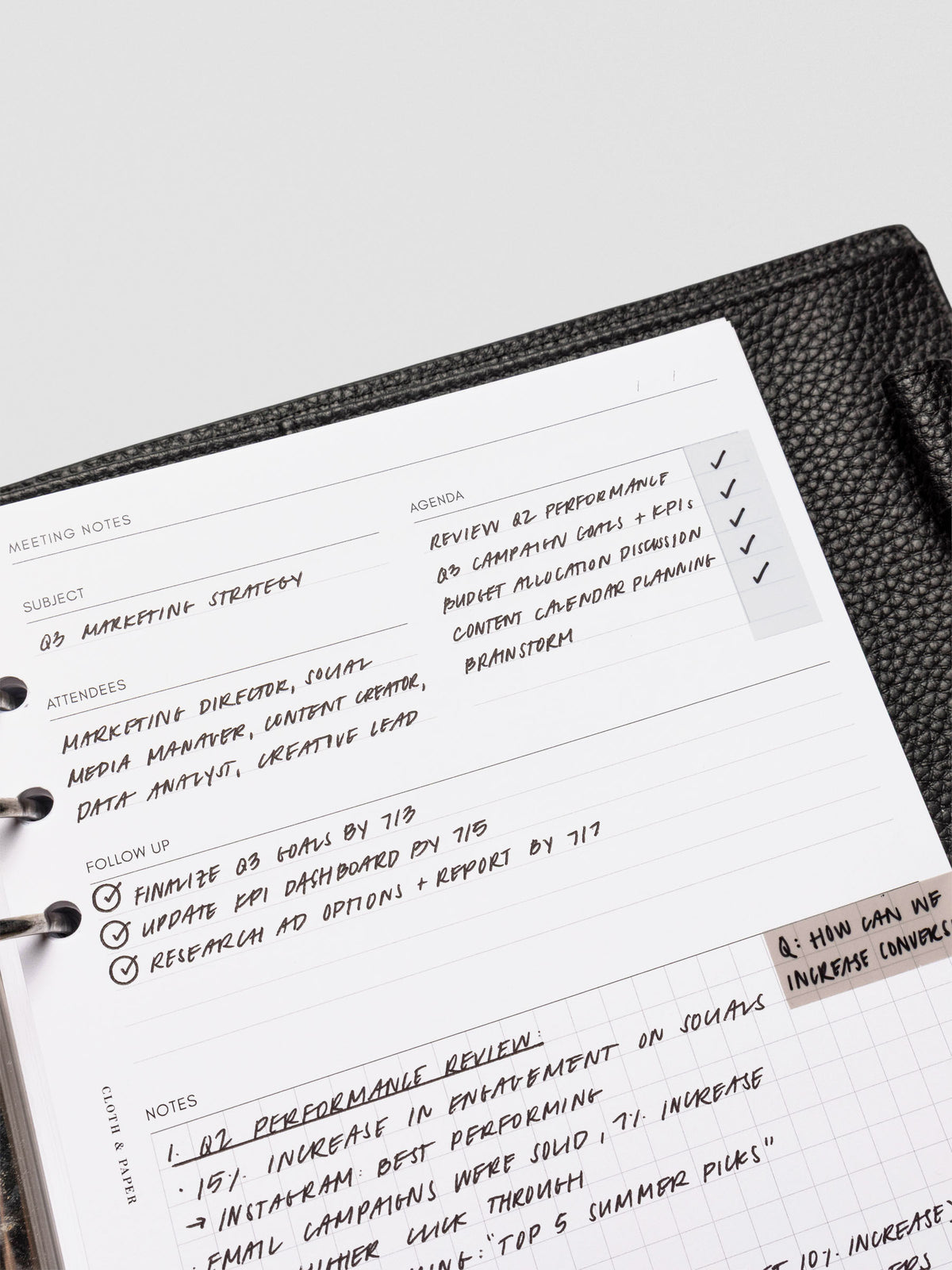A meeting notes insert filled in with handwritten text, featuring sections for subject, agenda, attendees, follow-up, and a gridded notes area. The page rests in a black leather planner.