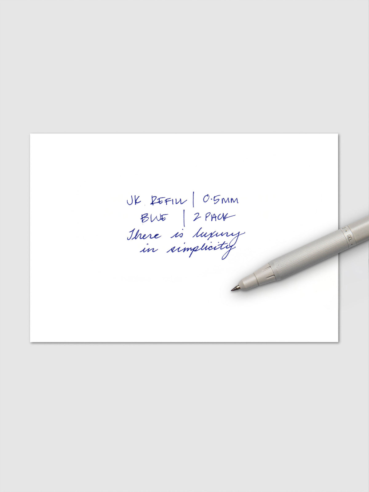 Close-up of handwritten text in blue ink reading "JK Refill | 0.5mm Blue | 2 Pack" followed by "There is luxury in simplicity," with a white Zebra pen resting on the paper.