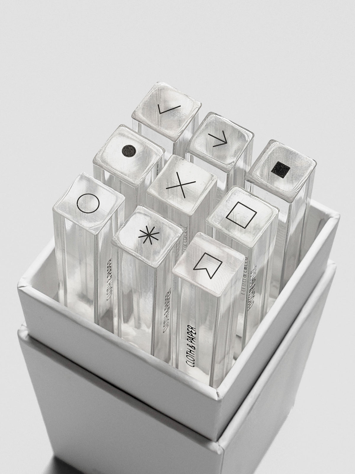 Open box revealing nine clear acrylic stamp sticks neatly arranged inside, each topped with a black icon.